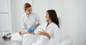 Portrait of charming woman sitting in armchair and holding glass of lemon water while doctor in sterile gloves checking IV infusion.