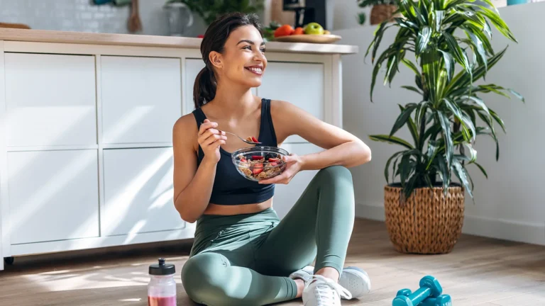 Athletic woman eating a healthy bowl of muesli with fruit sitting on floor in the kitchen at home